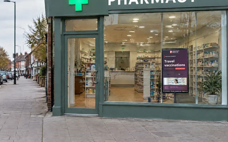 A brick-fronted pharmacy on a British high street with a portrait window screen showing a Knightsoft CMS layout: travel vaccinations promotion, clock, weather and news